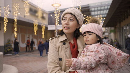 cheerful asian mother talking and holding her curious baby girl in arms while they are enjoying Christmas atmosphere outside a shopping mall with holiday lights