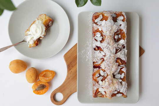 rectangular appetizing homemade apricot pie on a rectangular plate, on a wooden cutting board. Next to it is a round plate with a piece of cake and a spoonful of cream.