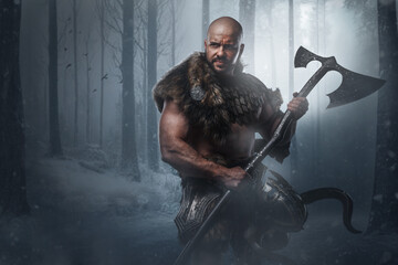 Portrait of nordic warrior with muscular build dressed in armor holding axe in winter wood.