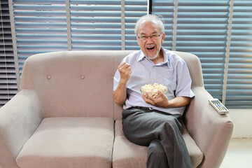 Happy smiling asian senior man sitting on sofa and eating popcorn while having fun with movie rest...