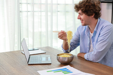 Young caucasian currly hair man sitting on table and eating tasty and instant noodle for lunch in...