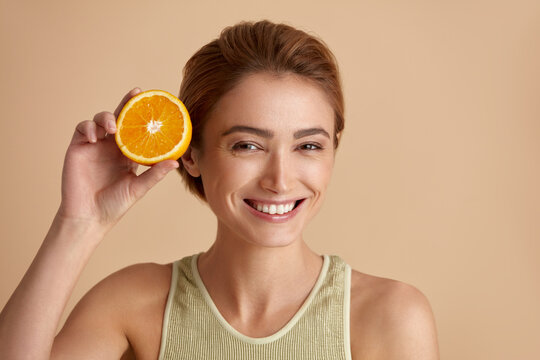 Laughing Girl Hold Slices of Orange. Happy Young Woman Looking at Camera and Recommended Vitamin for Skin. Concept of Skincare. Isolated on Beige Background 