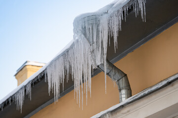 Big frozen icicles dangerously hanging from building edge on cold winter day, dangerous ice formation on metal house roof during bright sunny, but sub-freezing weather outside. Ice dam prevention - obrazy, fototapety, plakaty