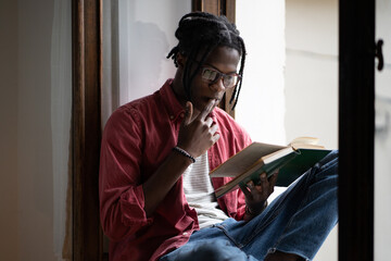 Young focused black man wearing eyeglasses learning foreign language by reading books, African American student guy sitting on windowsill at home Learning from textbook. College exams preparation - obrazy, fototapety, plakaty