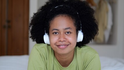 Obraz premium A young afro woman with black beautiful curly hair with a funny, calm and friendly mood spends her free leisure time lying down, listening to music wearing headphones on the bed a cozy room.