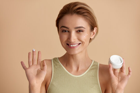 Caucasian Woman Applying Cream. Closeup Of Female Model With Fresh Skin Holding Cream Bottle In Hand. Portrait Of Happy Girl Applying Cosmetic Product Under Eyes. Skincare Concept. High Resolution 