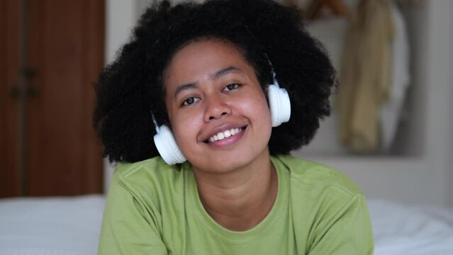 A young afro woman with black beautiful curly hair with a funny, calm and friendly mood spends her free leisure time lying down, listening to music wearing headphones on the bed a cozy room.