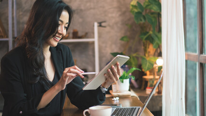 Professional businesswoman drinking coffee and working with clients using tablet and laptop on table in modern workspace