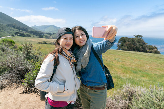 two smiling asian Taiwanese women tourists taking picture together with smartphone on beautiful landscape background at big sur California usa