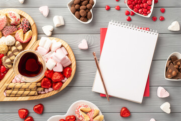 Valentine's Day concept. Top view photo of notepad wooden heart shaped serving tray glass of drinking saucers with confectionery candies chocolate cookies on grey wooden desk background with copyspace