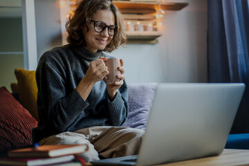 Young smiling woman in eyeglasses and warm sweater sitting on a sofa in front of a laptop with a mug of coffee and looking at the screen - obrazy, fototapety, plakaty