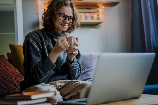 Young smiling woman in eyeglasses and warm sweater sitting on a sofa in front of a laptop with a mug of coffee and looking at the screen