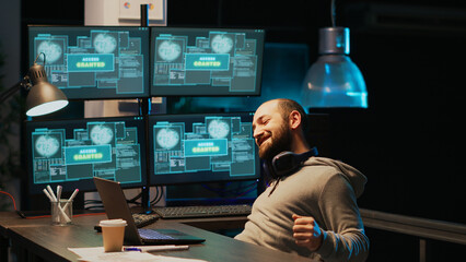 Cyber criminal celebrating hacking success in office at night, working with multiple monitors to steal password. Cheerful hacker with hidden identity feeling pleased about achievement.