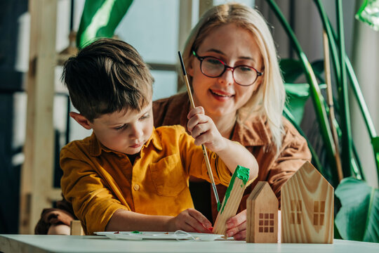 Smiling mother and son painting wooden model houses at table