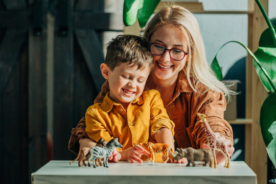 Happy mother and son playing with toy animal figurines on table at home