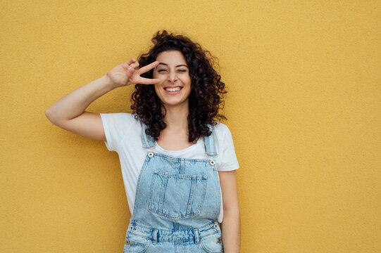 Happy woman winking and showing peace sign in front of yellow wall