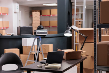 Empty large industrial warehouse equipped with tall metal shelves filled with various boxes and containers. Table desk full with shipping details document, laptop computer to work on delivery