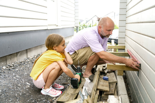 Father using leveling tool crouching with daughter near house