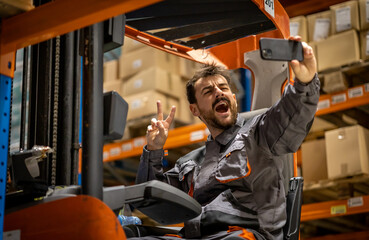 Young wearhouse forklift worker, having small break from work and using phone while sitting on...