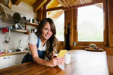 Smiling young woman leaning with smart phone on kitchen island