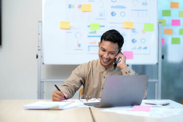 handsome young employee sitting at desk and looking at computer monitor while answering call in office