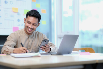 Fototapeta premium handsome young employee sitting at desk and looking at computer monitor while answering call in office