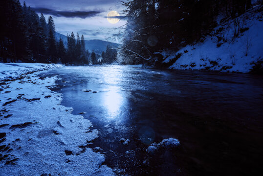 tereblya river in winter in full moon light. snow covered shore with coniferous forest on the shore. mountains beneath a cloudy sky on a frosty night in the distance. carpathian white season