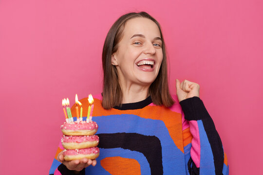 Image of cheerful extremely happy woman with brown hair wearing stylish jumper, clenched fist, rejoicing her birthday, holding donut with candles isolated over pink background.