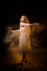 Fototapeta premium A girl in a white shirt and with long hair, looking like a ghost. A young model at a photo shoot with smoke, fog, flour, dust. A little fairy girl on a dark black background