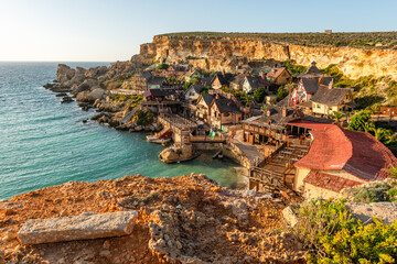 Fototapeta premium Malta, Il-Mellieha. View of the famous Popeye Village in Anchor Bay. Turquoise sea waters, blue sky and sunset light on cliffs