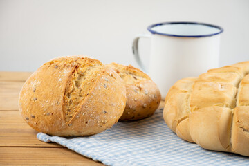 Close-up of loaf of rustic bread and rolls on cloth and wooden table with knife and cup, white background, horizontal, with copy space