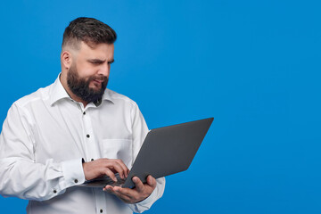 A bearded Caucasian man in a white shirt on a blue background holds a laptop. A solid man with a laptop.