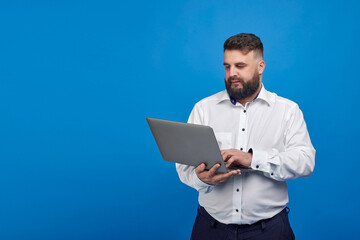 Naklejka na ściany i meble A bearded Caucasian man in a white shirt on a blue background holds a laptop. A solid man with a laptop.