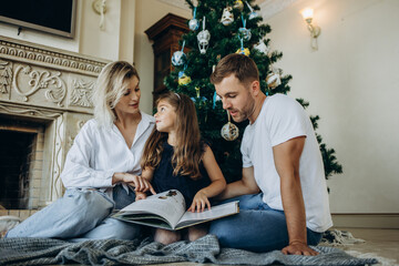 A daughter opens a gift next to her parents on the background of a Christmas tree