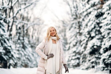 Fashion young smiling blonde woman in winter. Standing among snowy trees in winter forest. Wearing beige coat, white scarf in boho style