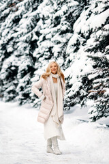 Fashion young smiling blonde woman in winter. Standing among snowy trees in winter forest. Wearing beige coat, white scarf in boho style