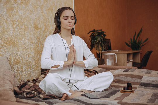 Female with closed eyes and Namaste hands practicing yoga and listening to meditation in earphones on bed in domestic room 