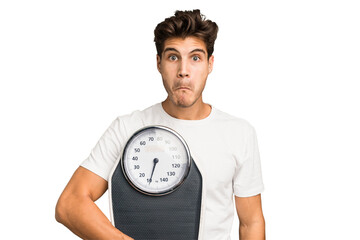 Young caucasian man holding a scale isolated shrugs shoulders and open eyes confused.
