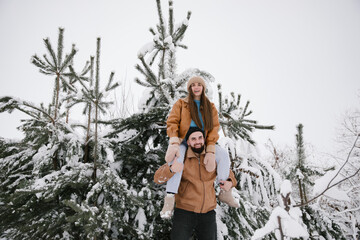 A woman sitting on the shoulders of a man in winter in the snow among snow-covered pines. A couple have fun in a snowy forest in winter.