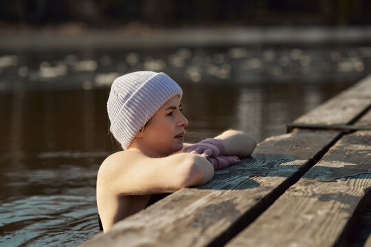 Caucasian adult woman swimming in frozen lake and leaning on wooden pier