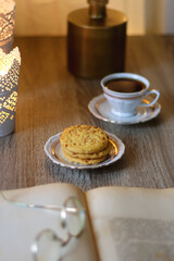 Plates with various cookies, cups of tea or coffee, vintage books, reading glasses and lit candles on the table. Selective focus.