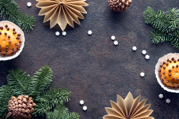 Wintertime dark background with fragrant pomander balls. Classical decorations - tangerine with cloves. Handmade paper stars from brown baking paper, pine cones, fir twigs and burning beewax candle.