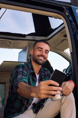 smiling young man taking a selfie with his mobile phone sitting in the door of his camper van, concept of freedom and digital nomad lifestyle