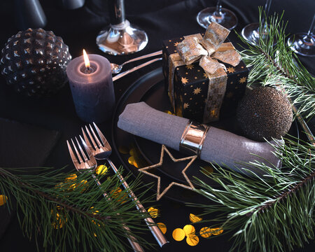 Atmospheric table decoration for a christmas or new year dinner. Place setting in modern style. Gift with golden loop and festive branch decoration, candle and golden confetti on black tablecloth.