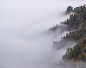 Mountain ranges with rocks overgrown with trees descend into clouds and fog, into a white shroud, a damp landscape in the autumn mountains