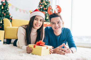 Young happy Asian woman wearing a Santa Claus hat with her boyfriend with a Christmas gift while lying down on the carpet with a Christmas tree in the background. Photo with copy space.