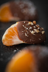 Orange juicy tangerine slices covered with melted milk chocolate on black concrete background. Sweet snack. Macro shot. Selective focus. Bokeh background