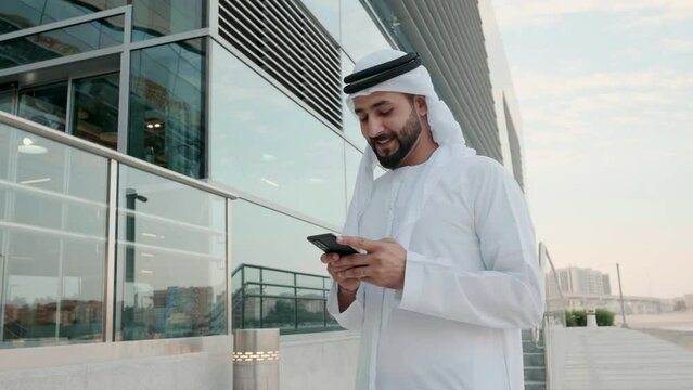 Happy Arab man in Kandoora using cell phone device Arabic Emirati local at a business location with glass building as background