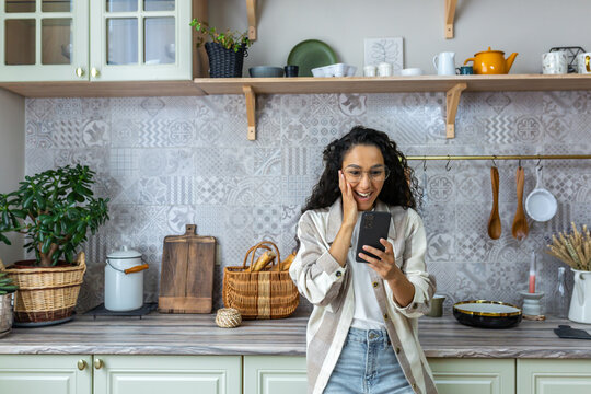 A Happy Latin American Woman Is Standing In The Kitchen At Home, Holding The Phone In One Hand, Holding Her Head With The Other Hand. I Am Glad, I Received An Unexpected And Joyful Message, Good News.