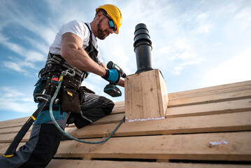 A carpenter with a safety harness and tool belt holds a electric screwdriver in his hand. He is...